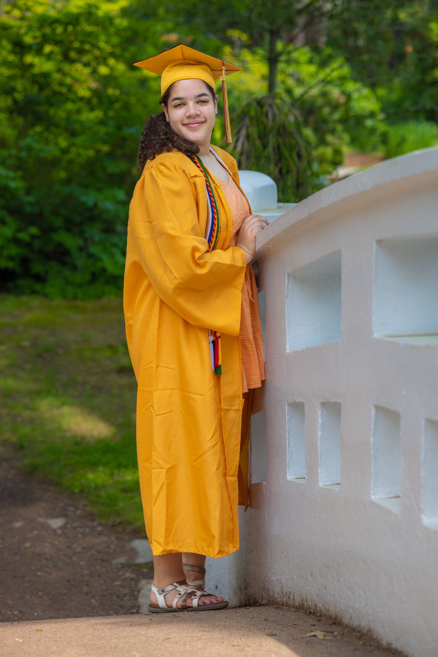 Graduate in bright yellow cap and gown poses on white bridge railing in garden setting.