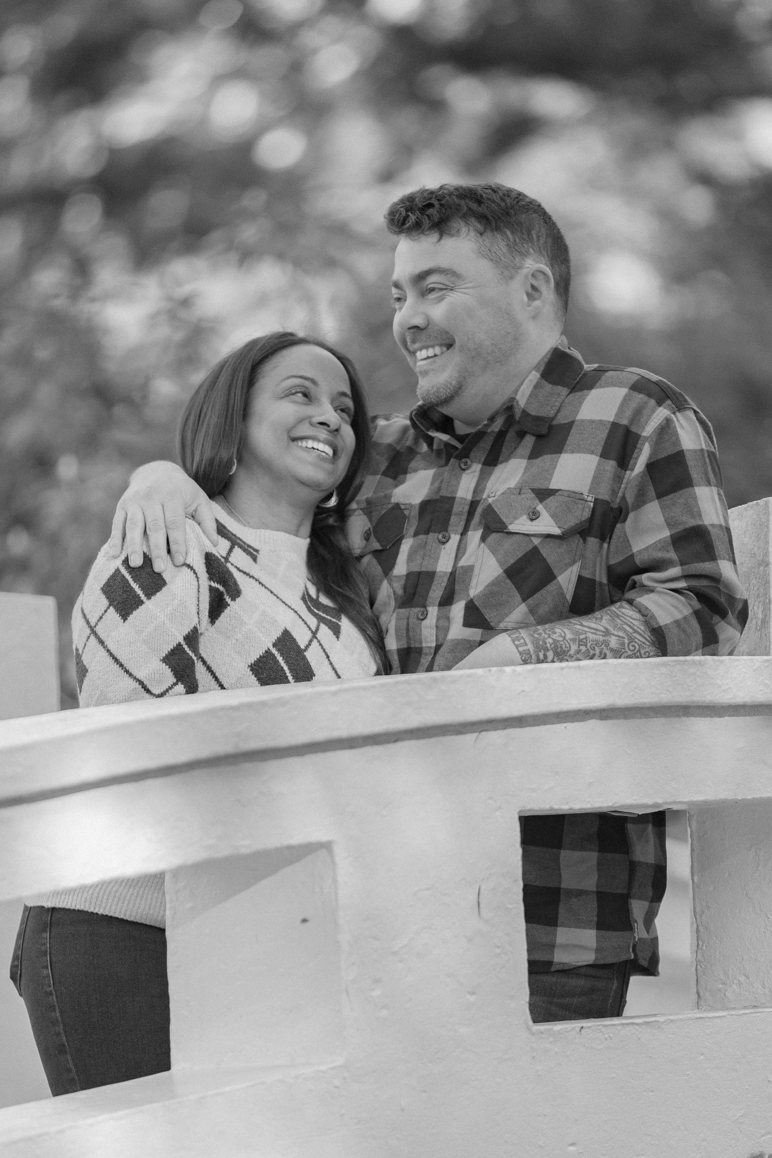 A loving couple shares a joyful moment together while leaning on a white railing outdoors in black and white.