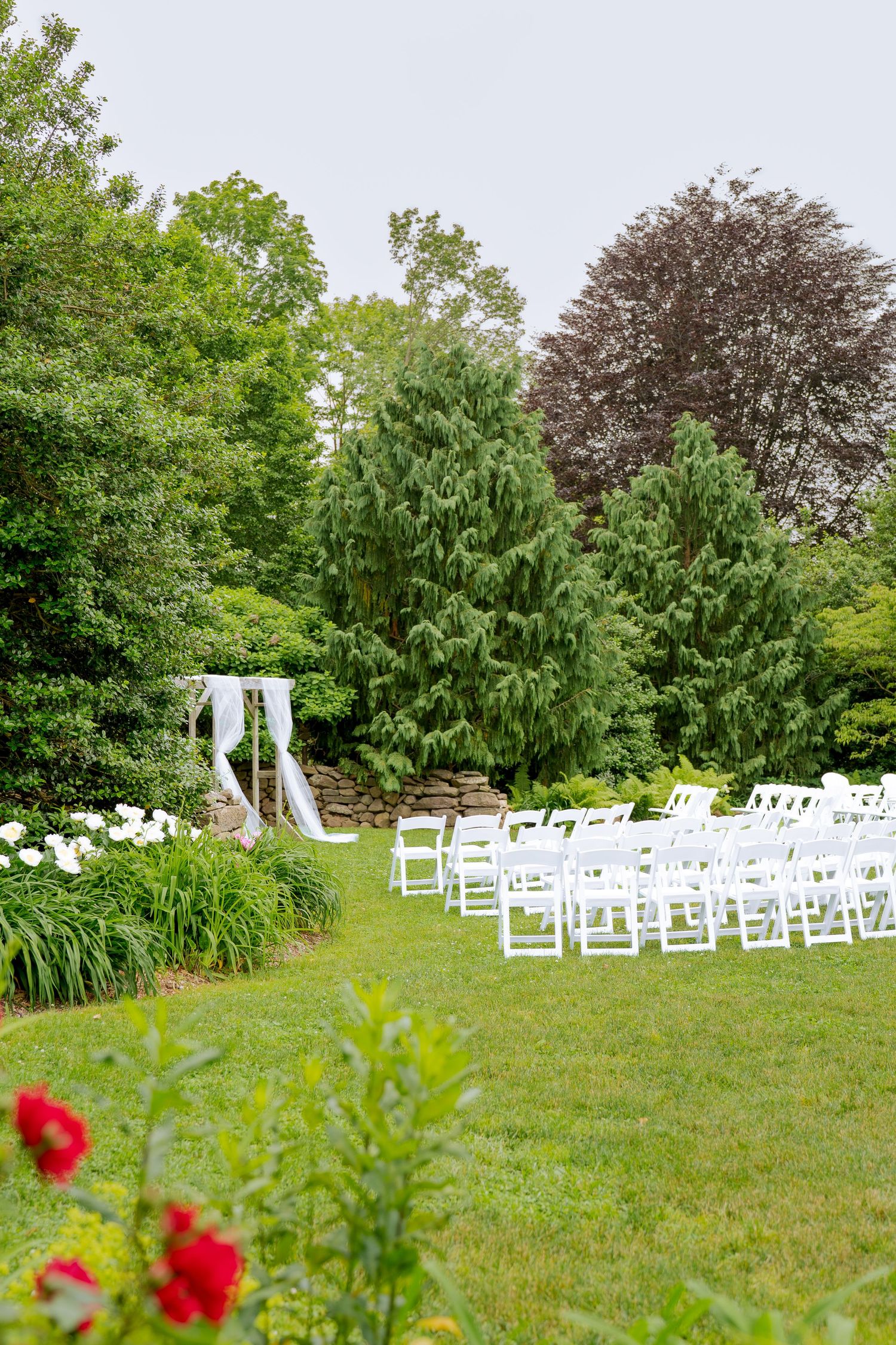 White wedding ceremony setup on a lush green lawn with an arch and chairs surrounded by evergreen trees in a garden setting.