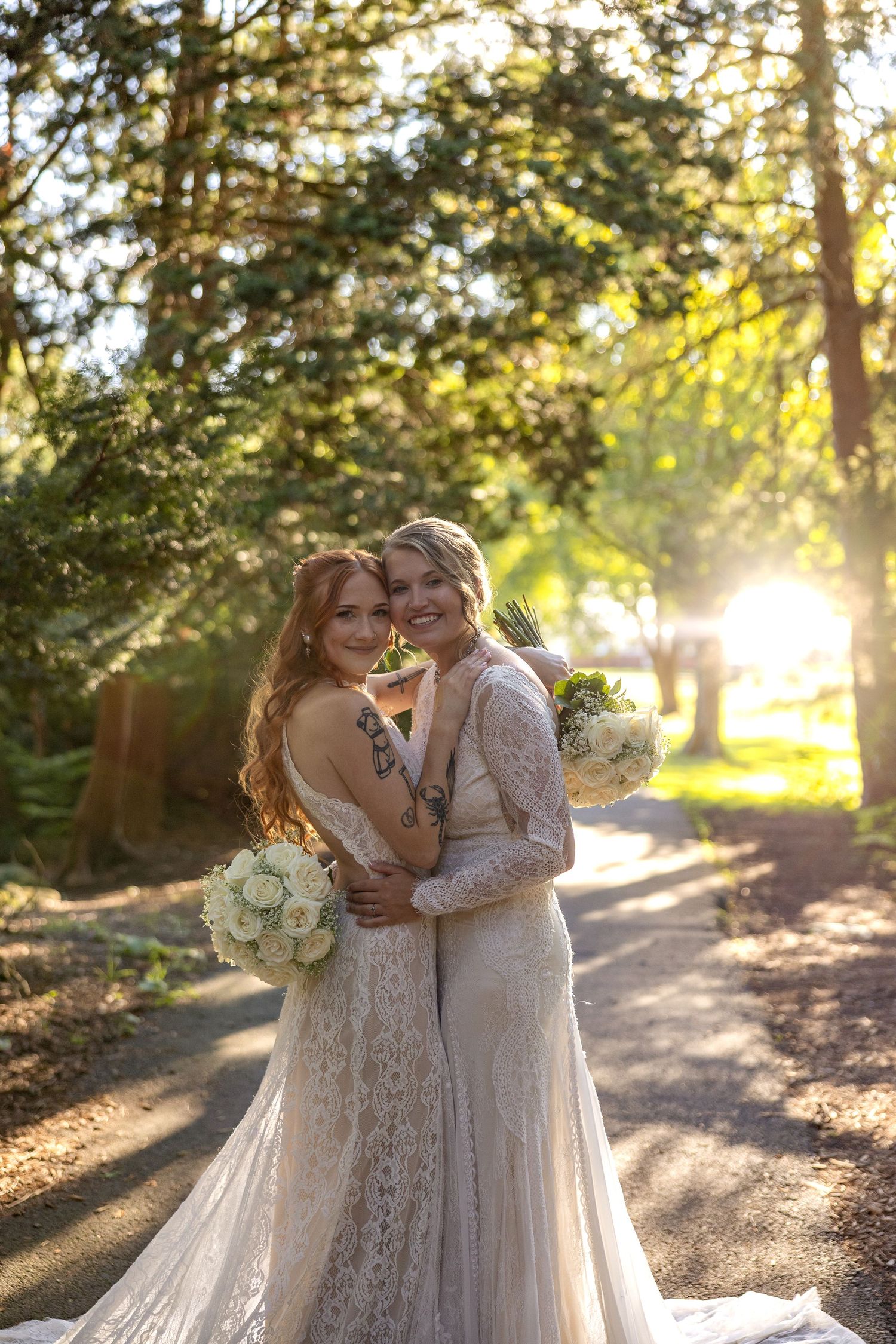 A couple in elegant white dresses share an intimate moment in a sunlit forest path during golden hour.