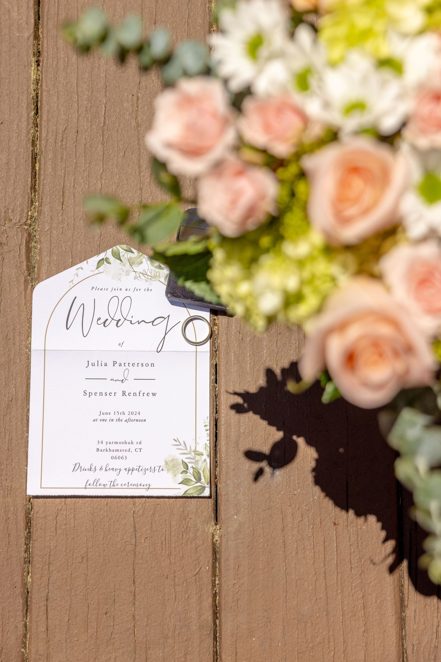 Wedding invitation with pink roses and greenery creating shadows on a wooden surface.