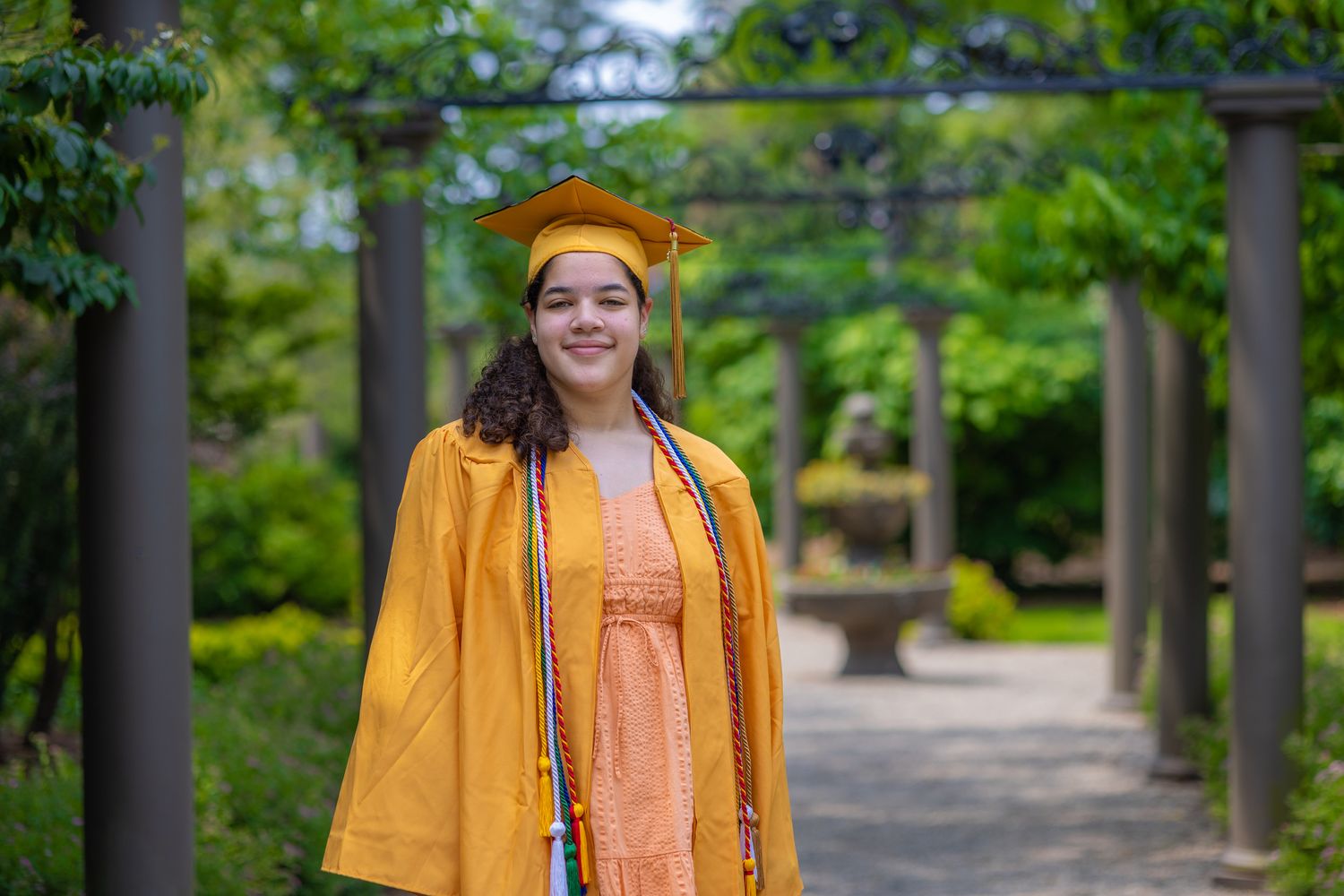 Graduate in gold regalia stands smiling in a garden archway.
