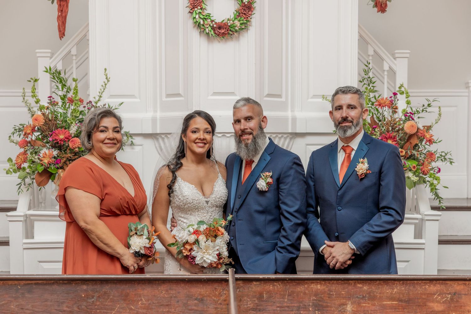 Wedding party poses in front of white church doors decorated with coral floral arrangements.