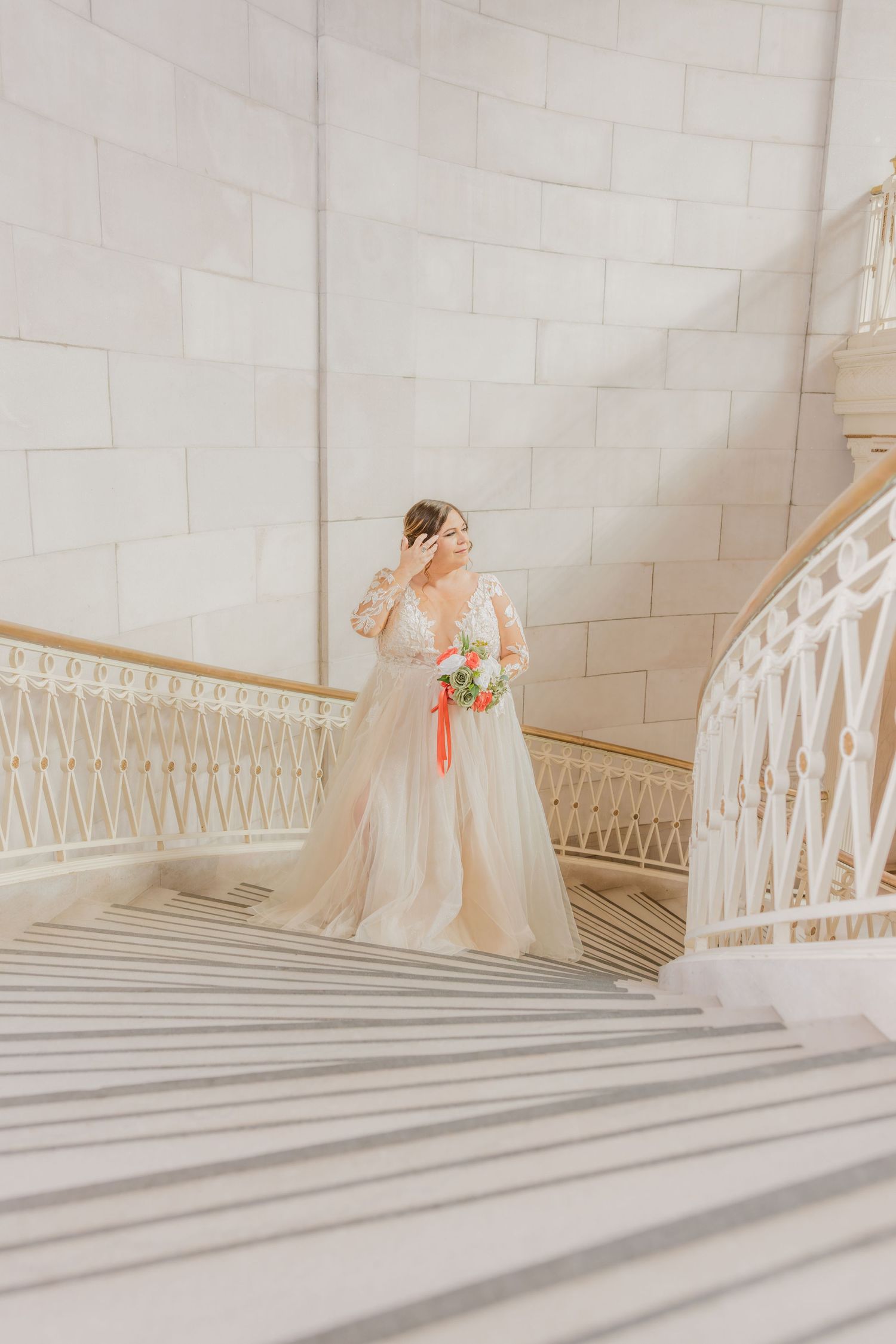 Bride poses on elegant white marble staircase holding coral bouquet.