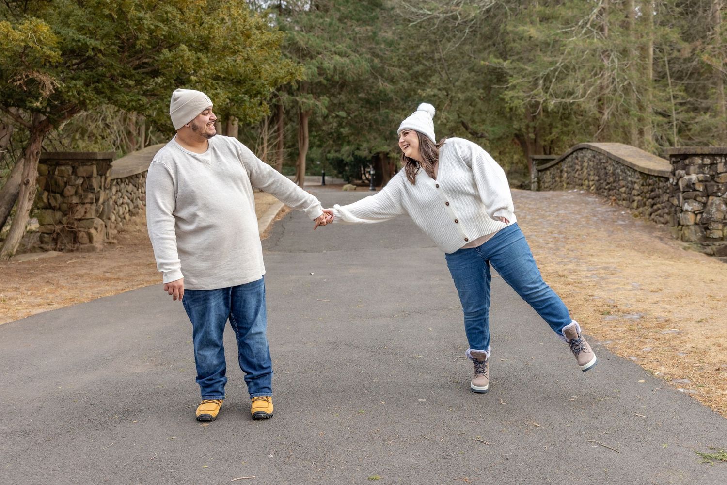 Playful couple in matching sweaters hold hands while dancing on a paved path.