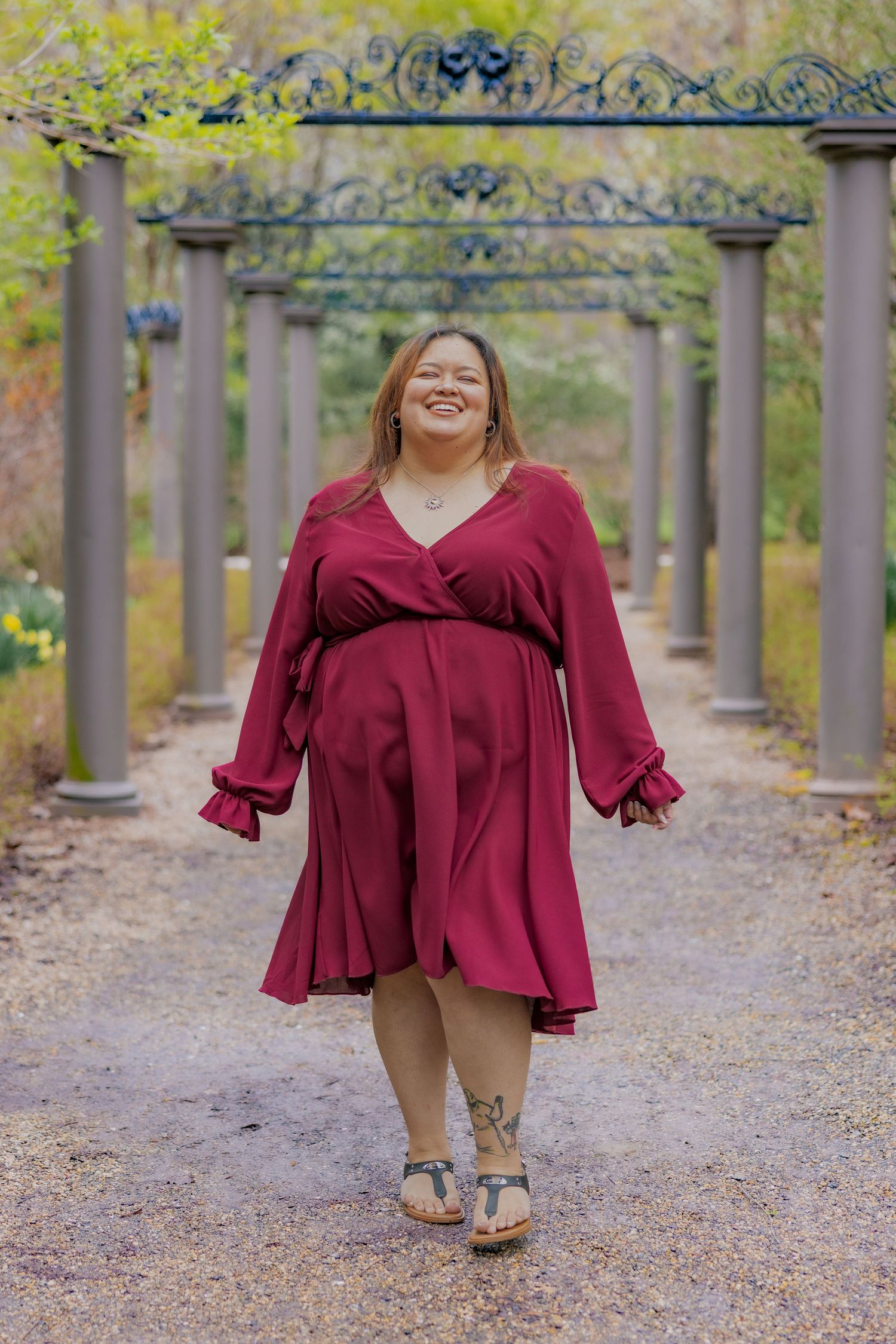 Person wearing burgundy dress poses joyfully under ornate garden trellis archway on gravel path.
