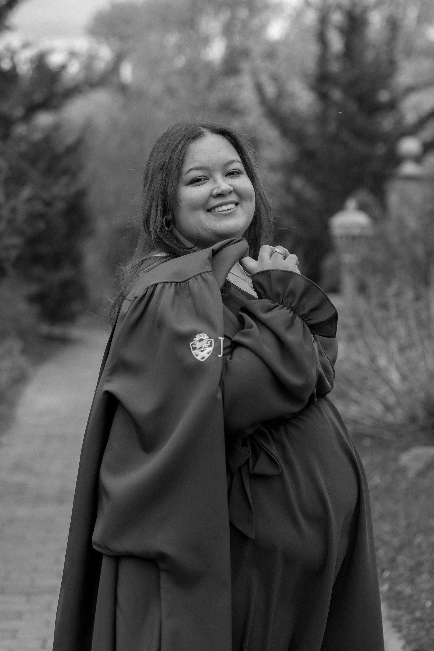 Graduate in black academic regalia poses outdoors in black and white portrait style.