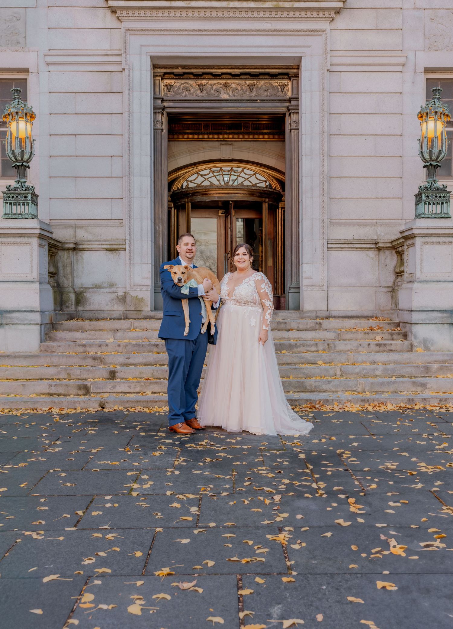 Wedding couple poses in front of elegant building entrance with autumn leaves scattered on ground.