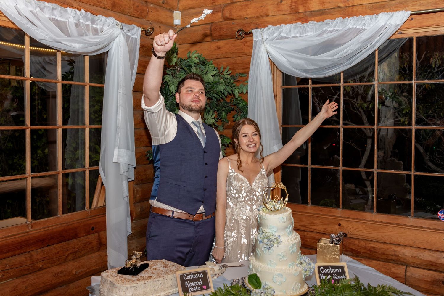 Newlyweds celebrate with raised arms next to their elegant white wedding cake in a rustic wooden venue.