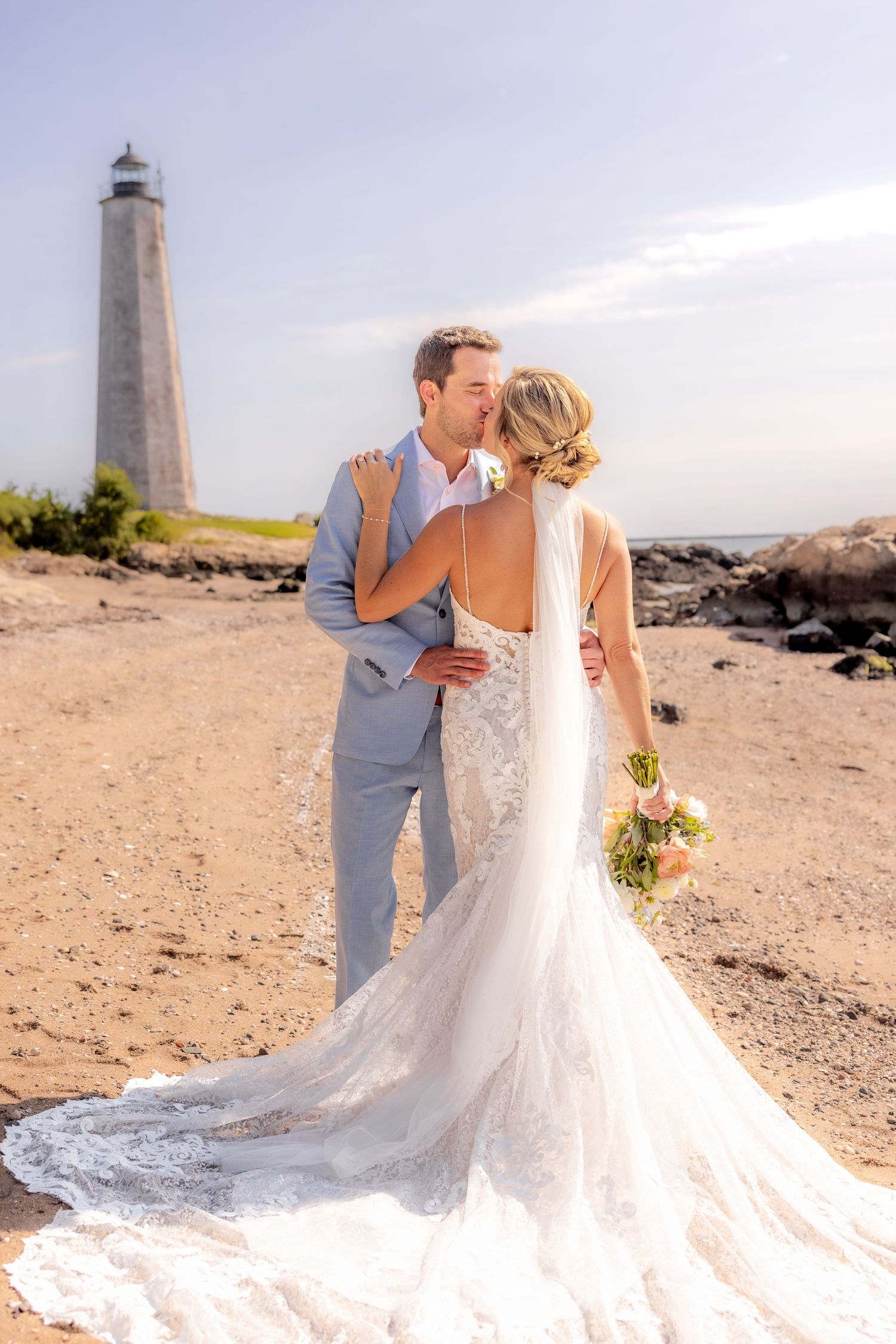 Wedding couple poses romantically by a lighthouse on a rocky beach with flowing white dress train.