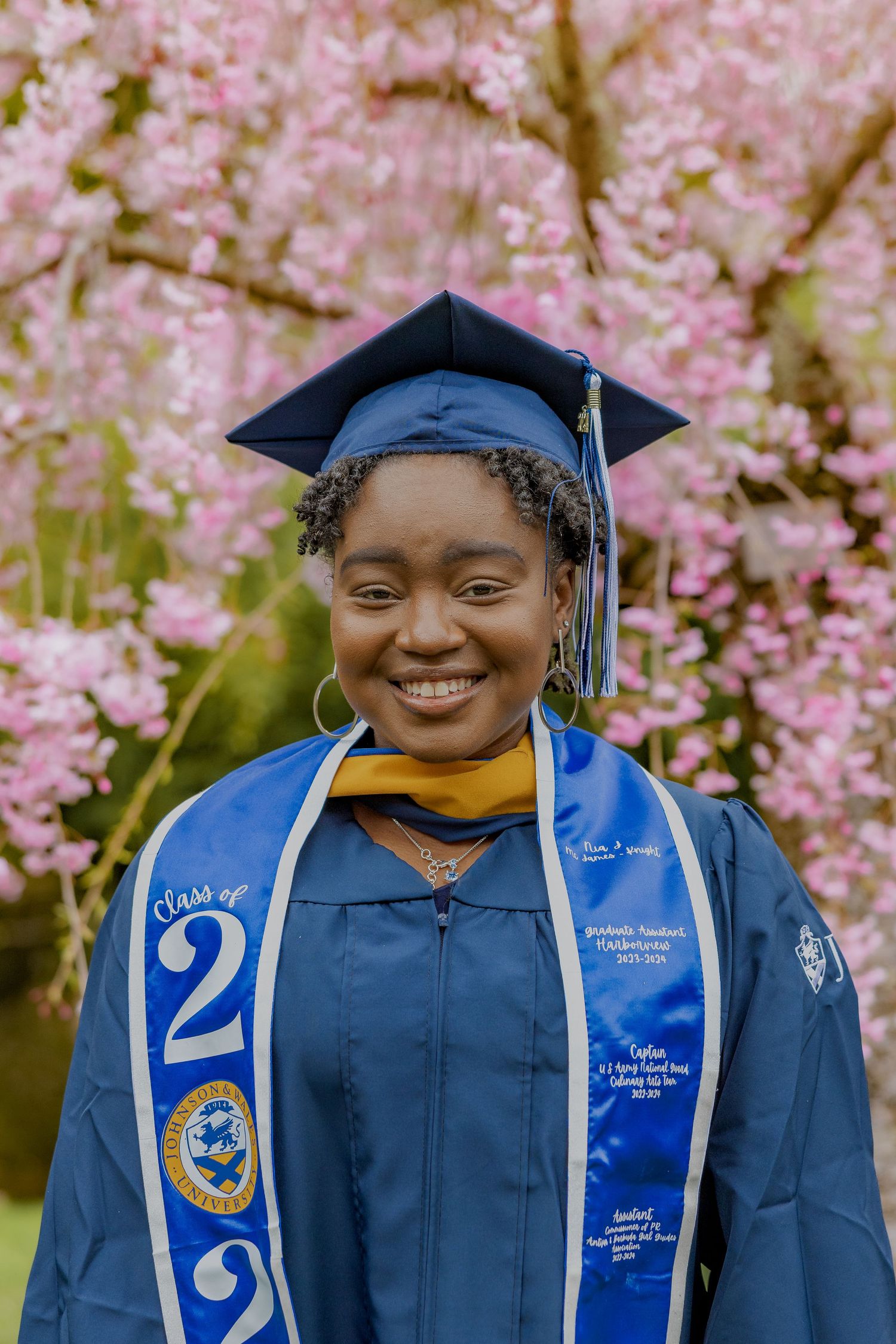 A graduate in blue cap and gown with a Class of 2022 sash poses in front of blooming cherry blossoms on a spring day.