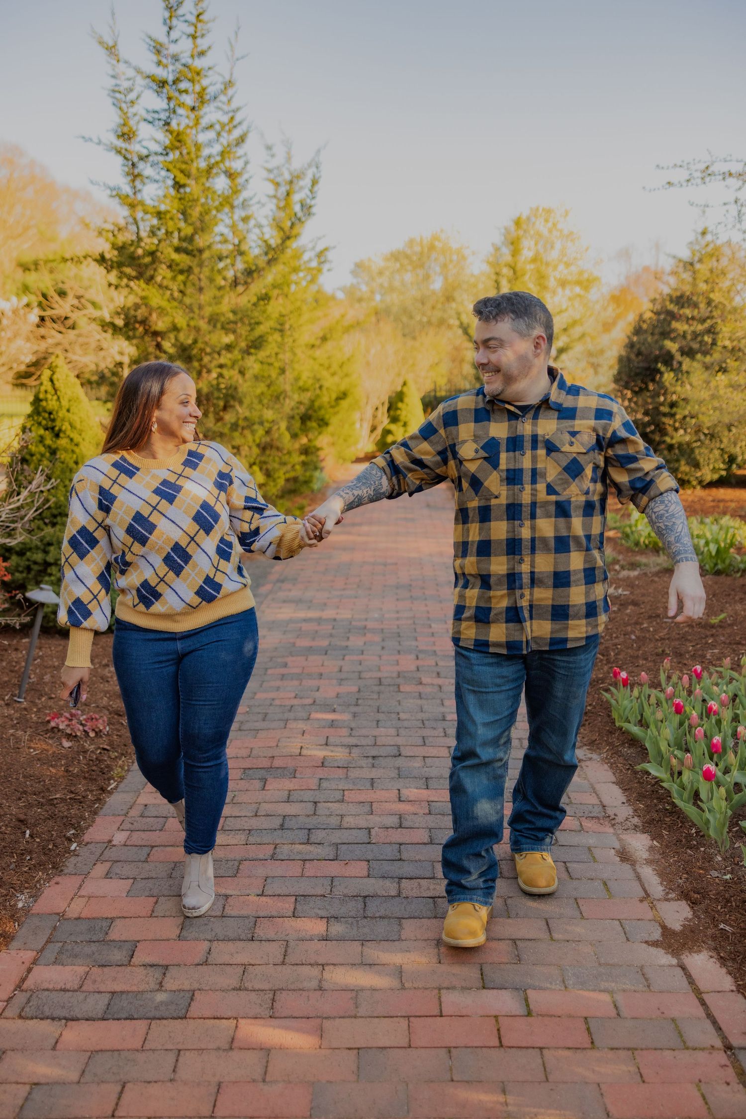 A couple wearing matching plaid shirts walk together on a brick path lined with tulips and evergreen trees.