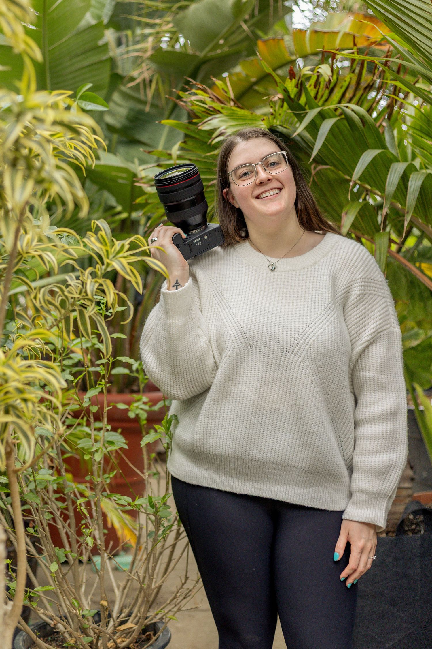Photographer holding professional camera poses among tropical plants in greenhouse setting.