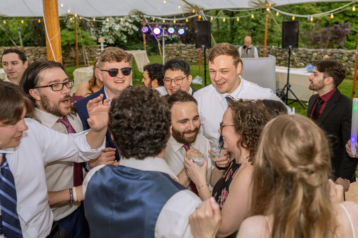 A lively crowd of wedding guests gather in a circle on an outdoor dance floor under string lights during a reception celebration.