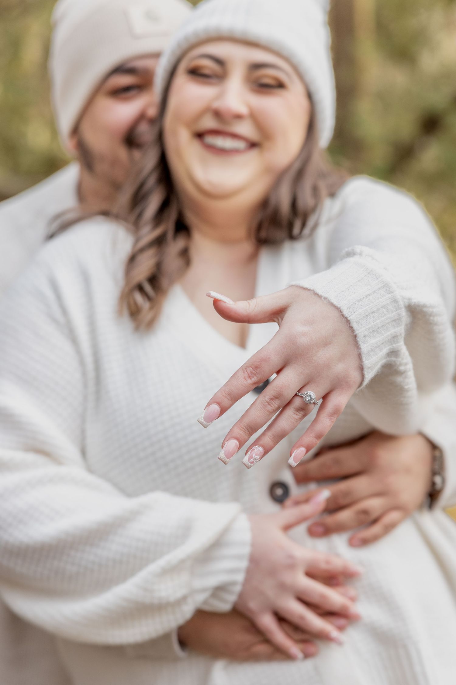 Close-up of an engagement ring being shown while couple embraces in white winter sweaters.
