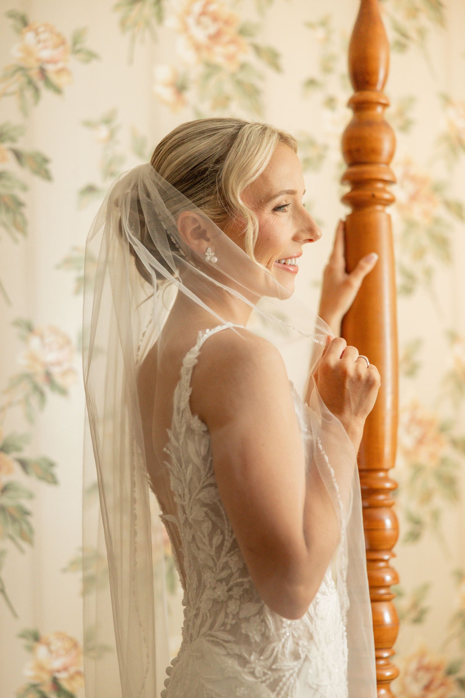 Side profile of bride in lace dress with veil against floral wallpaper.