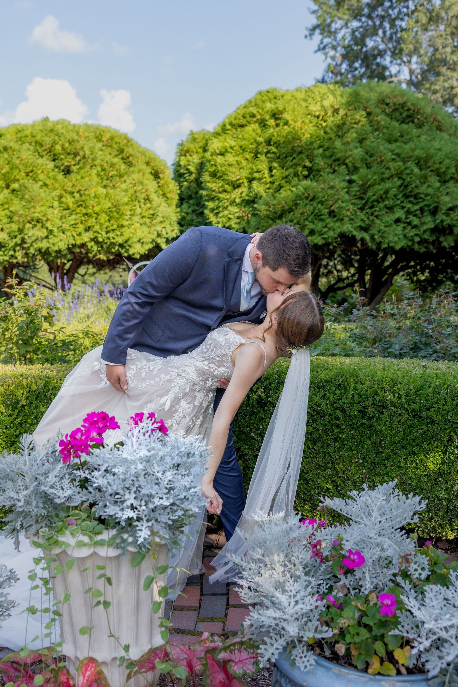 Romantic wedding couple sharing a kiss near blooming pink flowers and hedges.