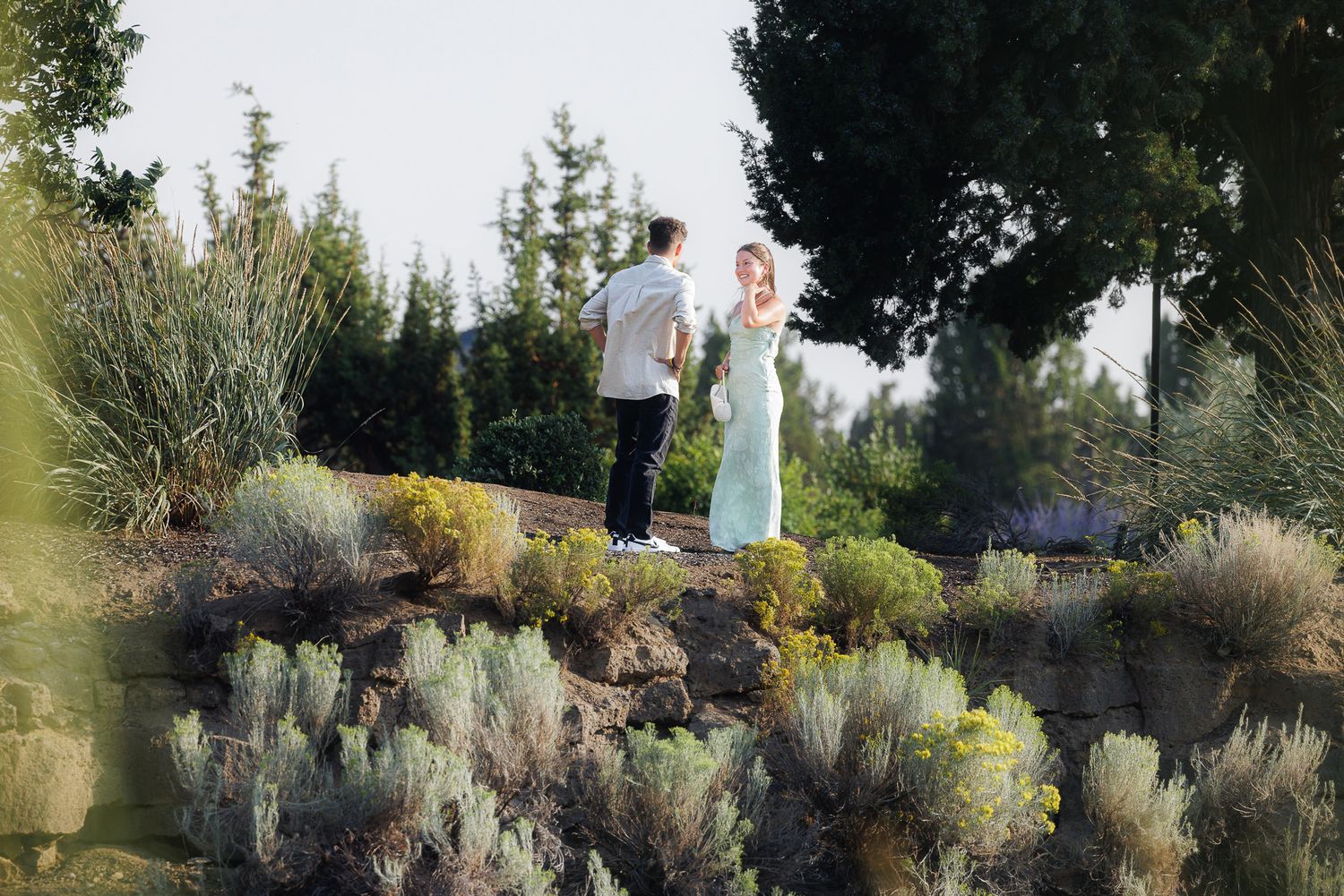 Couple standing on a rocky hillside surrounded by lush greenery and lavender plants in a scenic outdoor garden.
