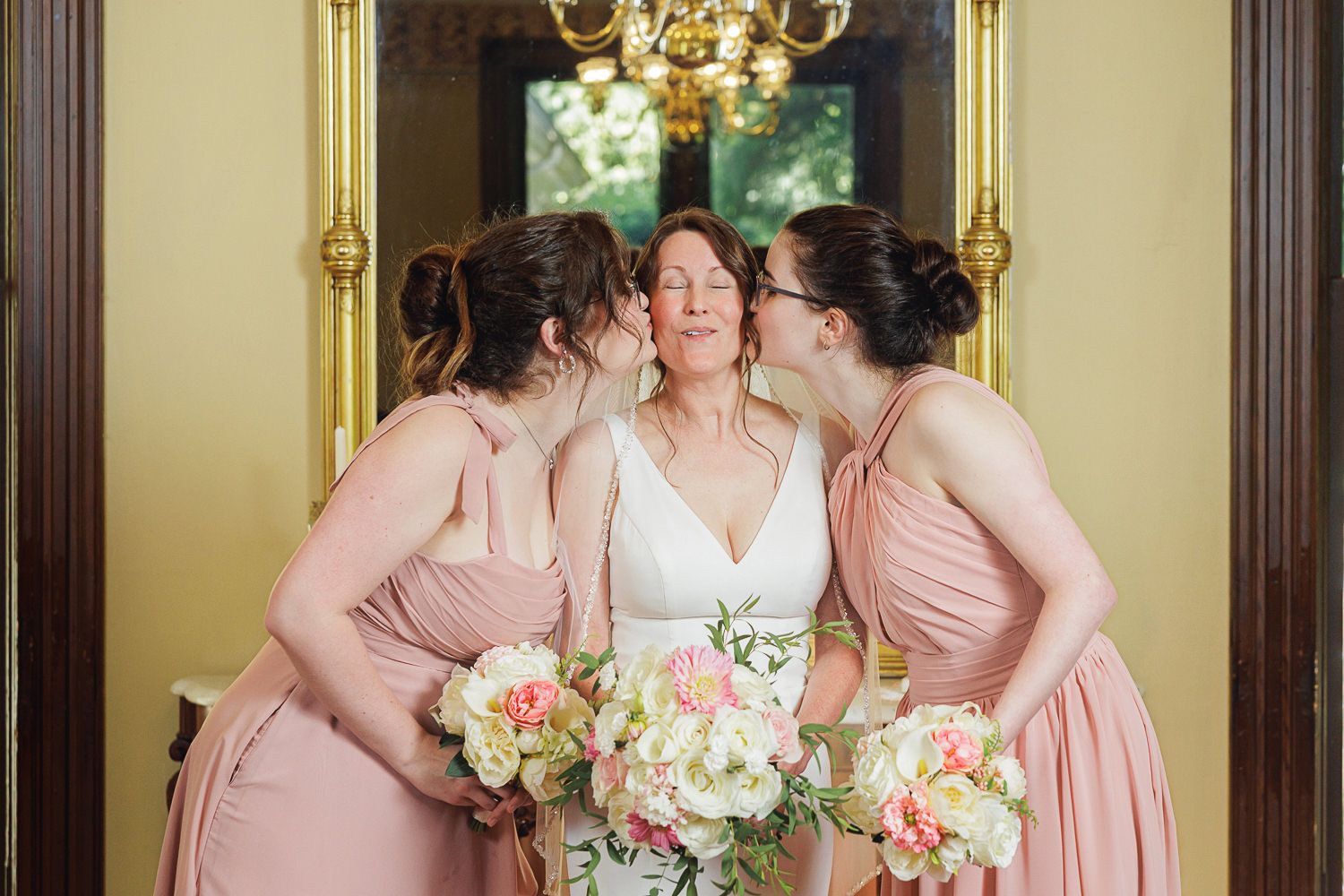 A wedding party in pink and white dresses poses together holding pink and white bouquets in an elegant indoor setting.