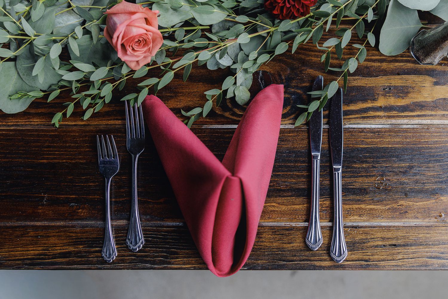 A rustic wooden table setting with burgundy napkins and silver cutlery arranged in a row against dark wood.