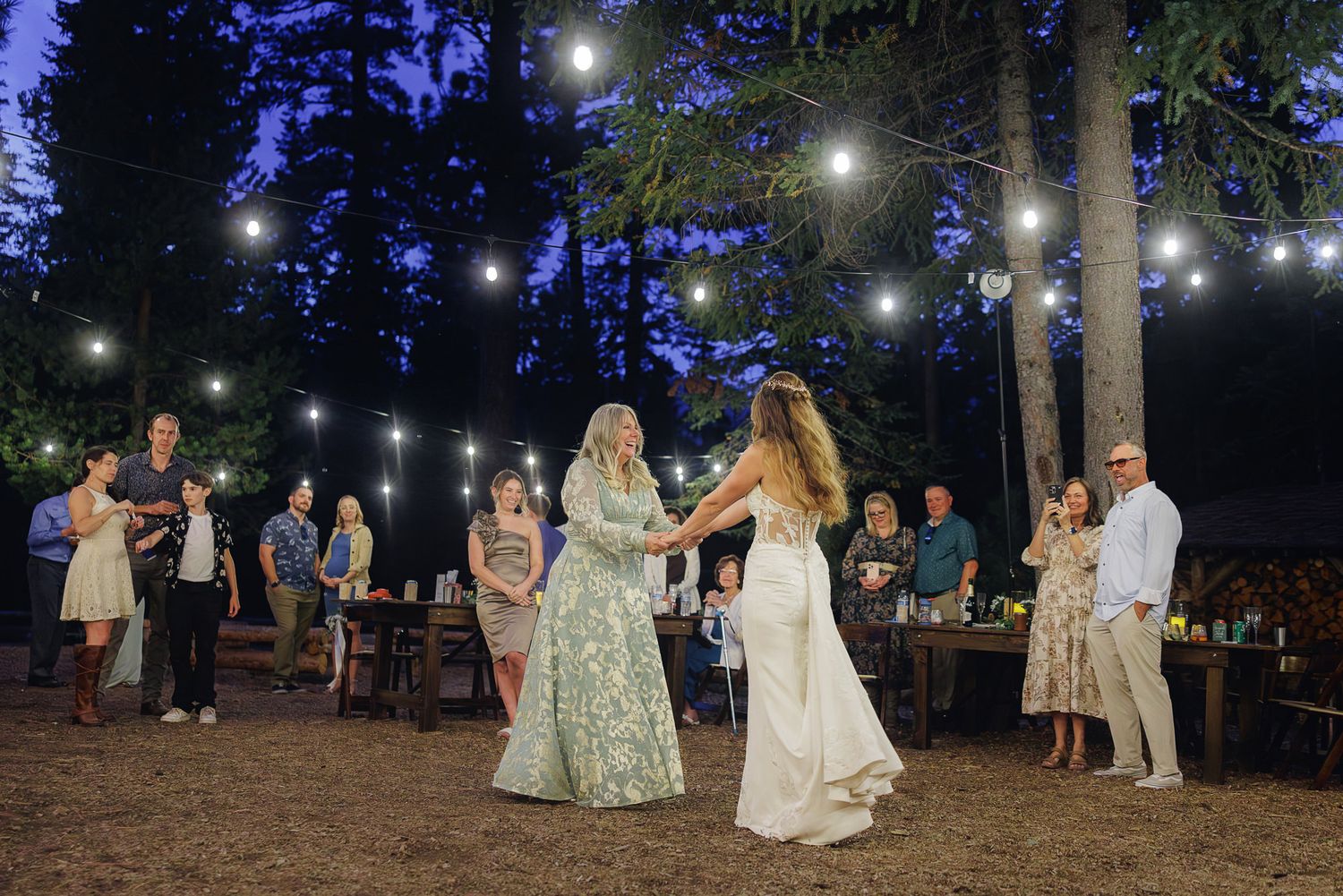 A romantic outdoor wedding reception in the woods with string lights illuminating couples dancing under tall trees.