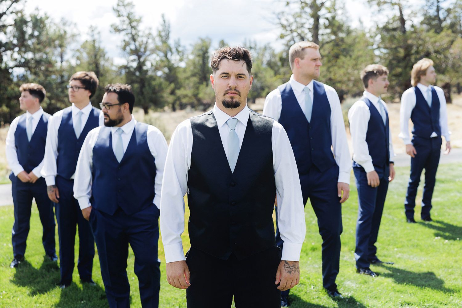 A wedding party wearing blue vests and white shirts poses on a sunny lawn with pine trees in the background.