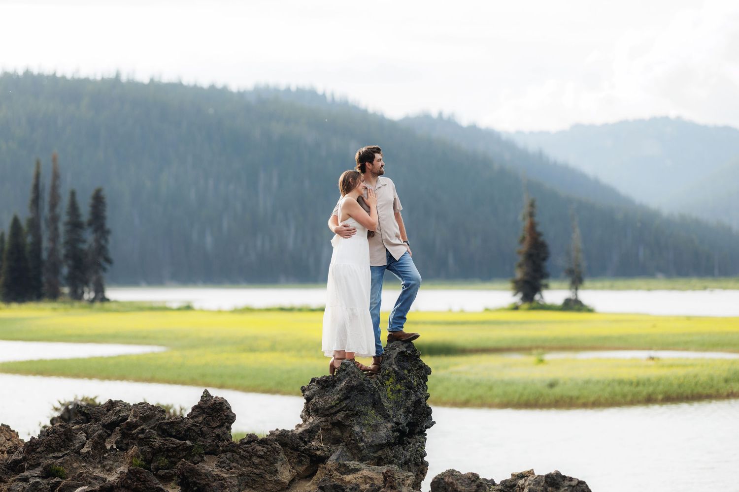 A couple in wedding attire poses on a hilltop overlooking mountains and a tranquil lake on their special day.