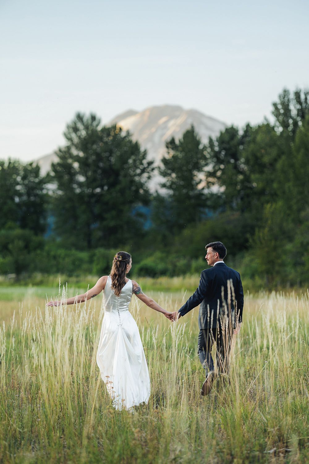 A couple in wedding attire walks hand in hand through a tall grass meadow with mountains in the background.