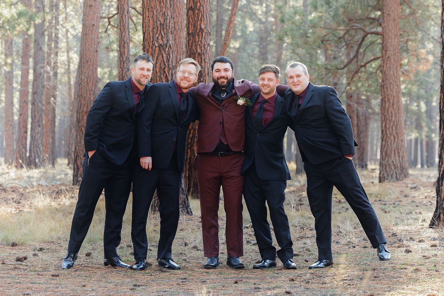 A group of five people in dark formal attire pose together outdoors in a pine forest setting.