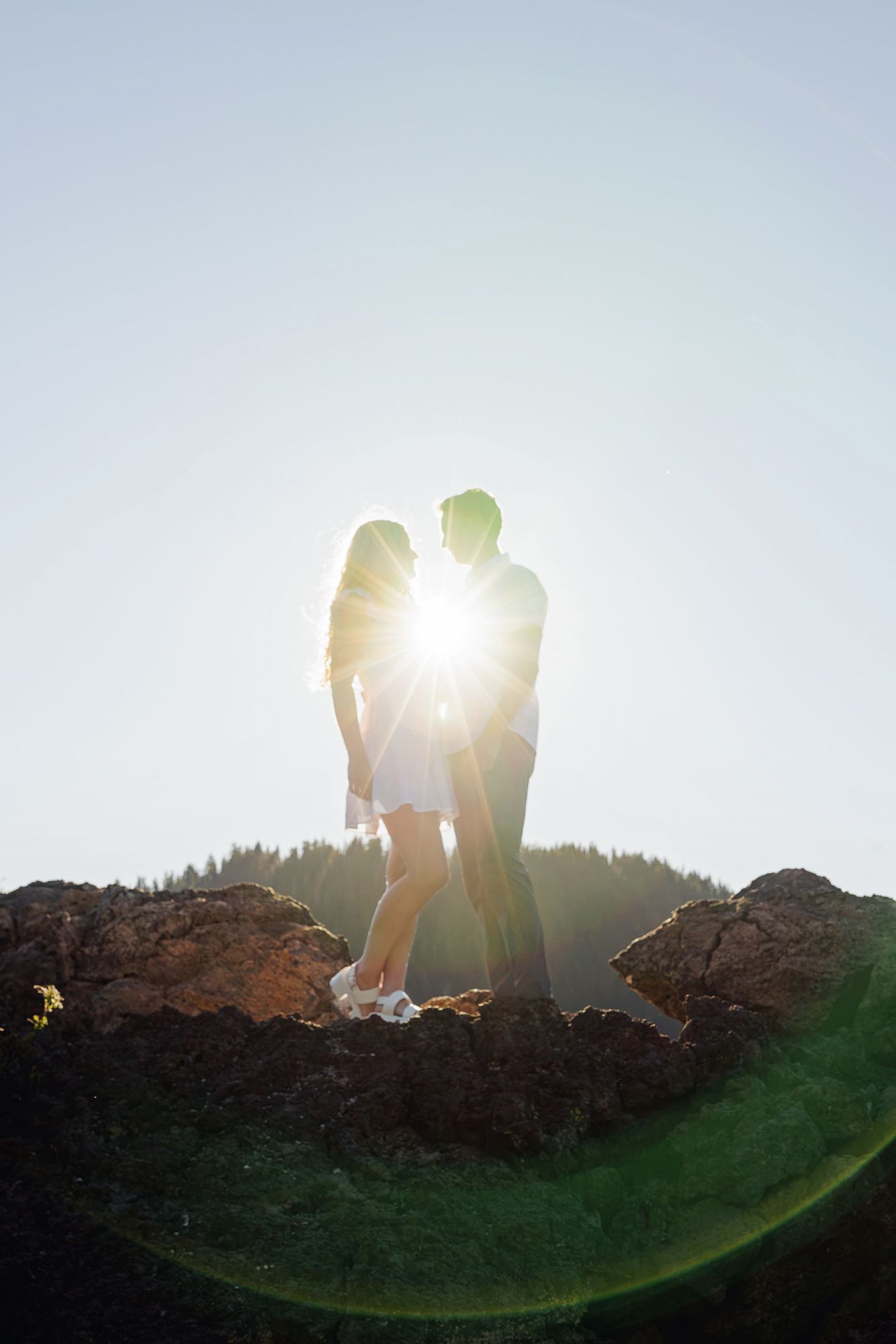 Couple silhouetted against bright sunlight standing on rocky hilltop with green grass, romantic outdoor portrait.