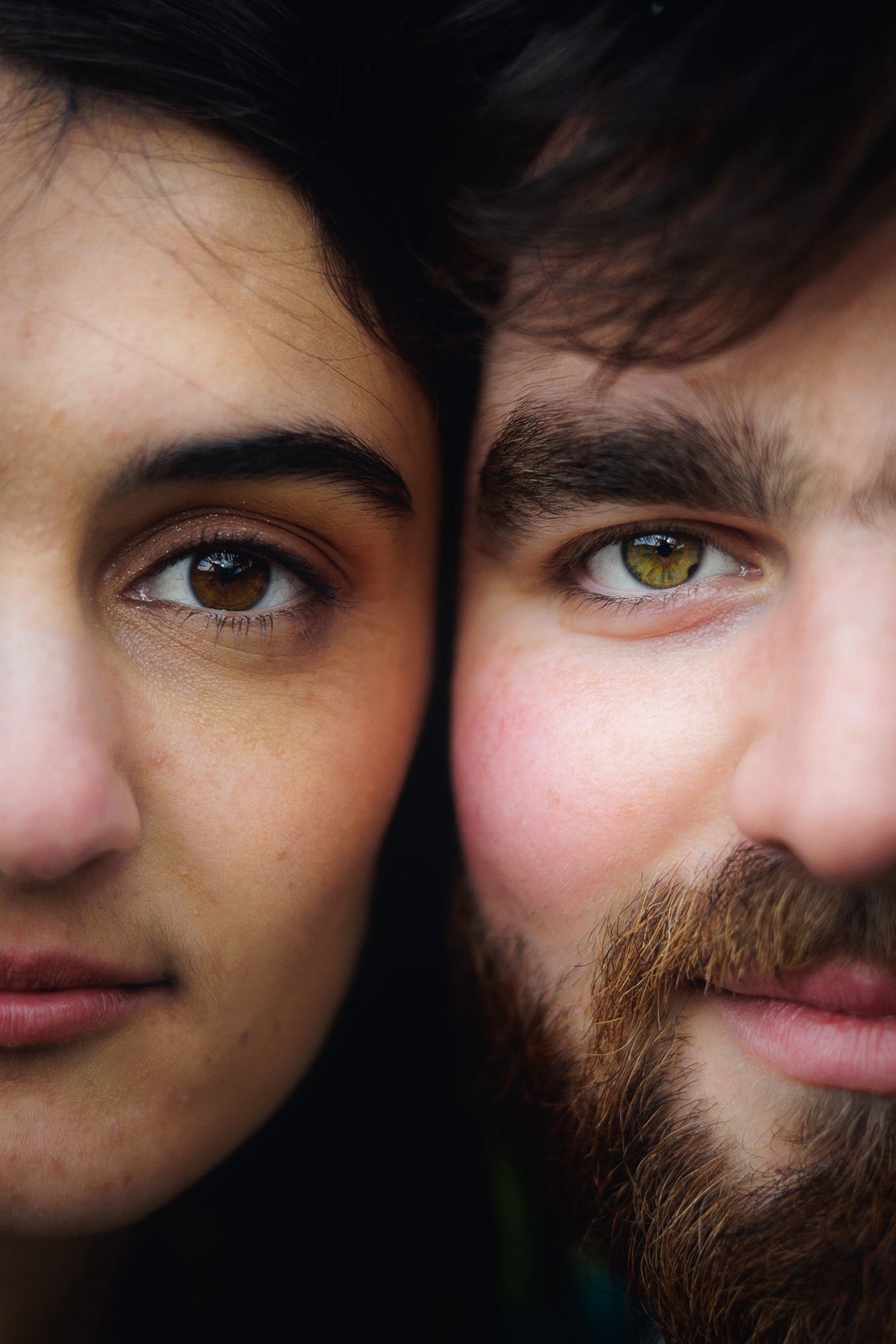 Close-up portrait of a woman and bearded man with striking hazel eyes side by side against dark background.
