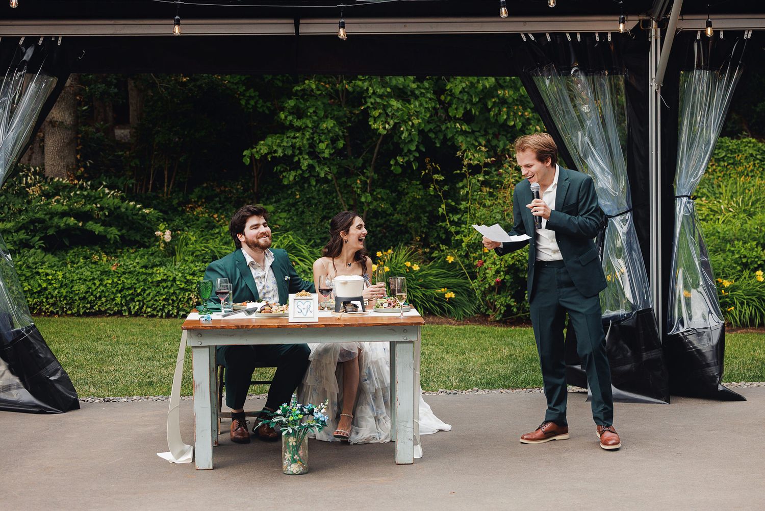 Groom reads wedding speech at outdoor reception while bride and groom sit at decorated sweetheart table.
