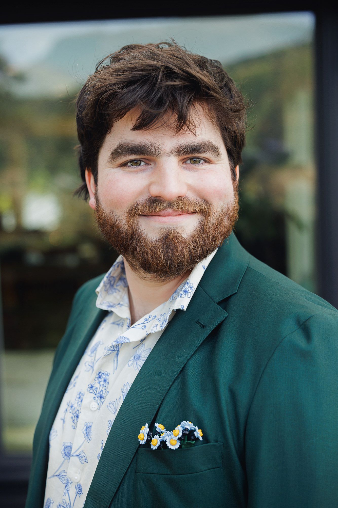Young man with beard wearing green blazer and floral shirt with daisy pocket square, smiling outdoors.