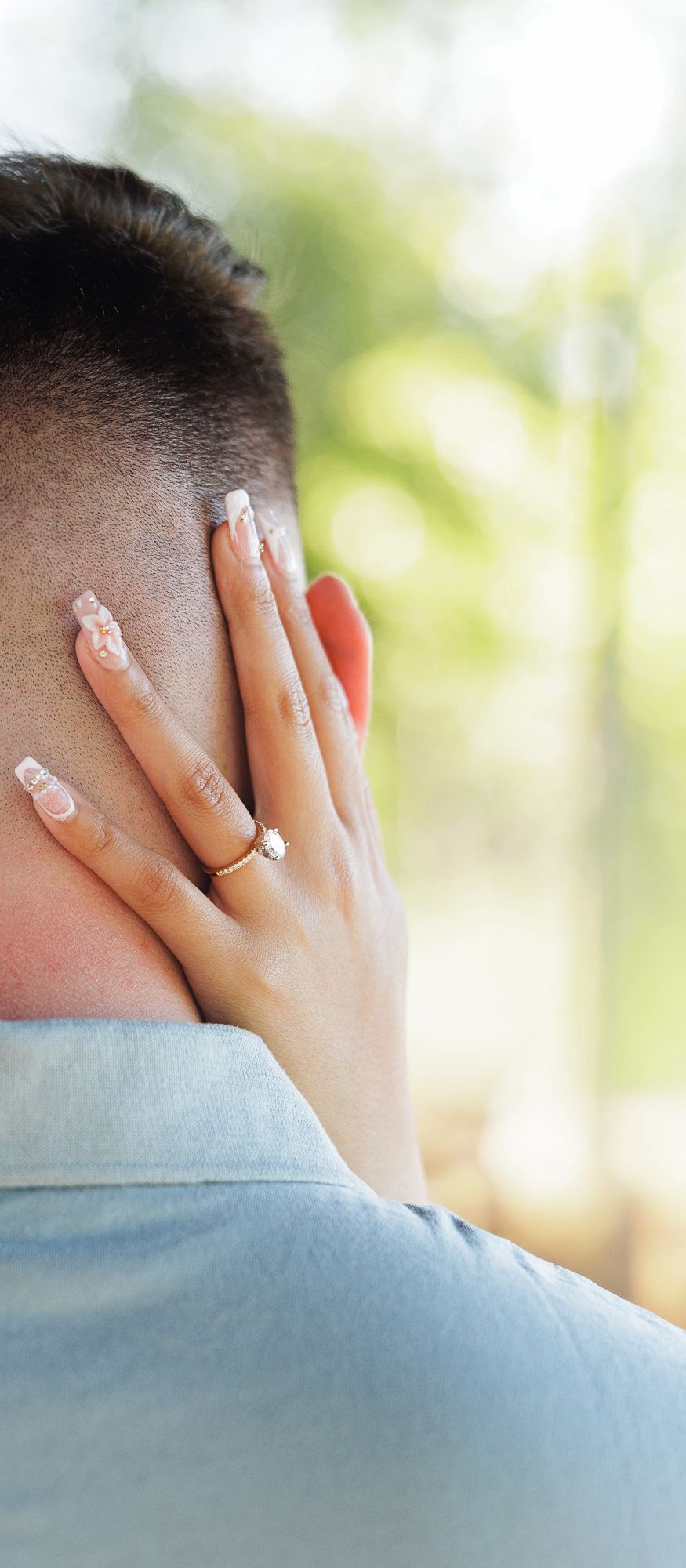 Close-up of woman's hand with diamond engagement ring resting on man's neck outdoors.