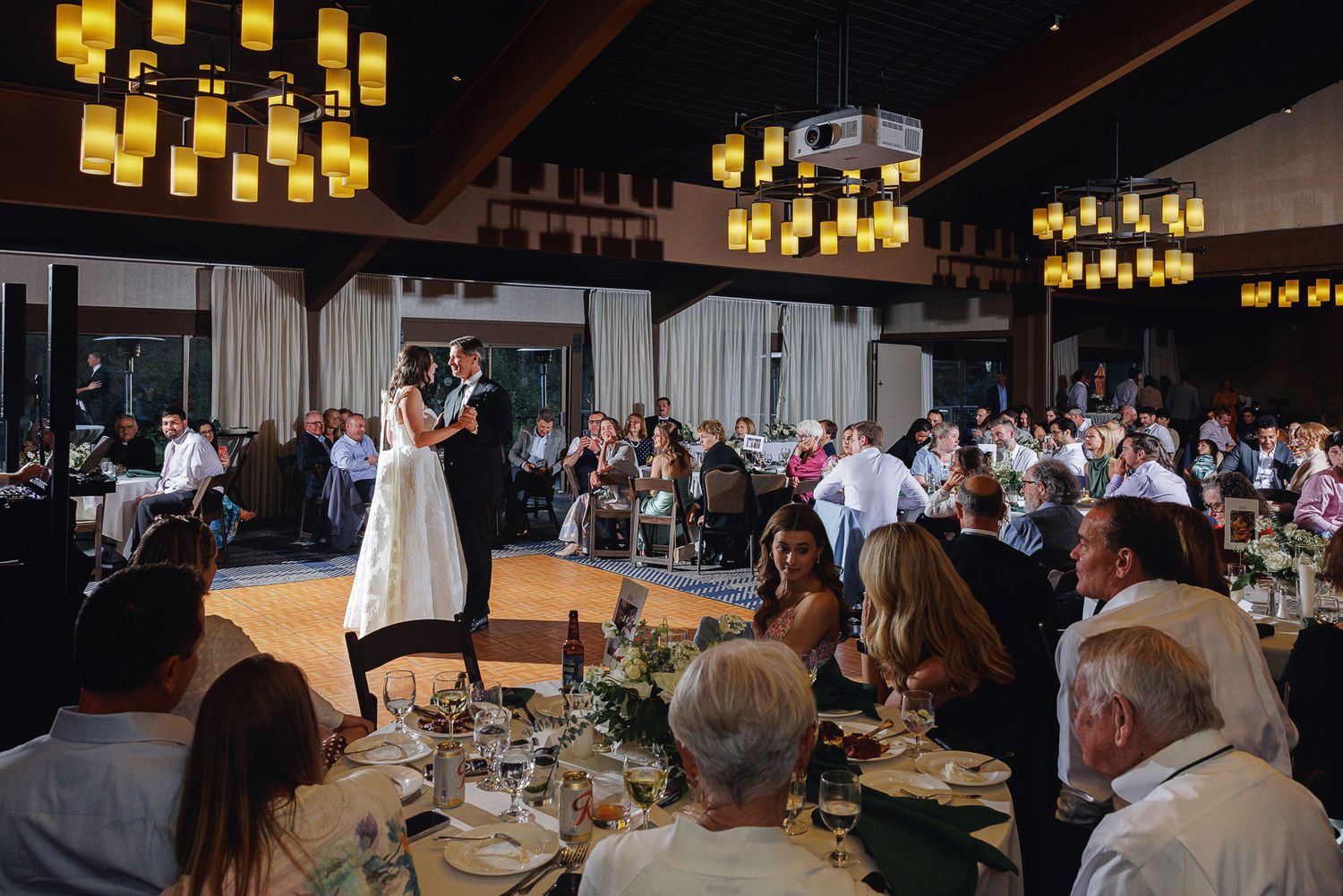 Bride and groom share first dance at elegant wedding reception with warm chandelier lighting and seated guests.