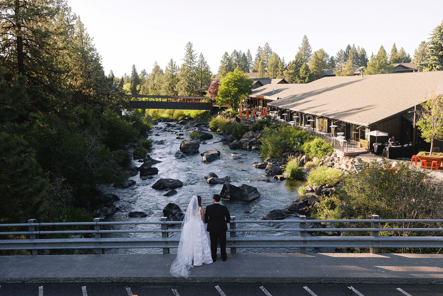 Bride and groom stand on a bridge overlooking a scenic river surrounded by tall pine trees at their outdoor wedding venue.