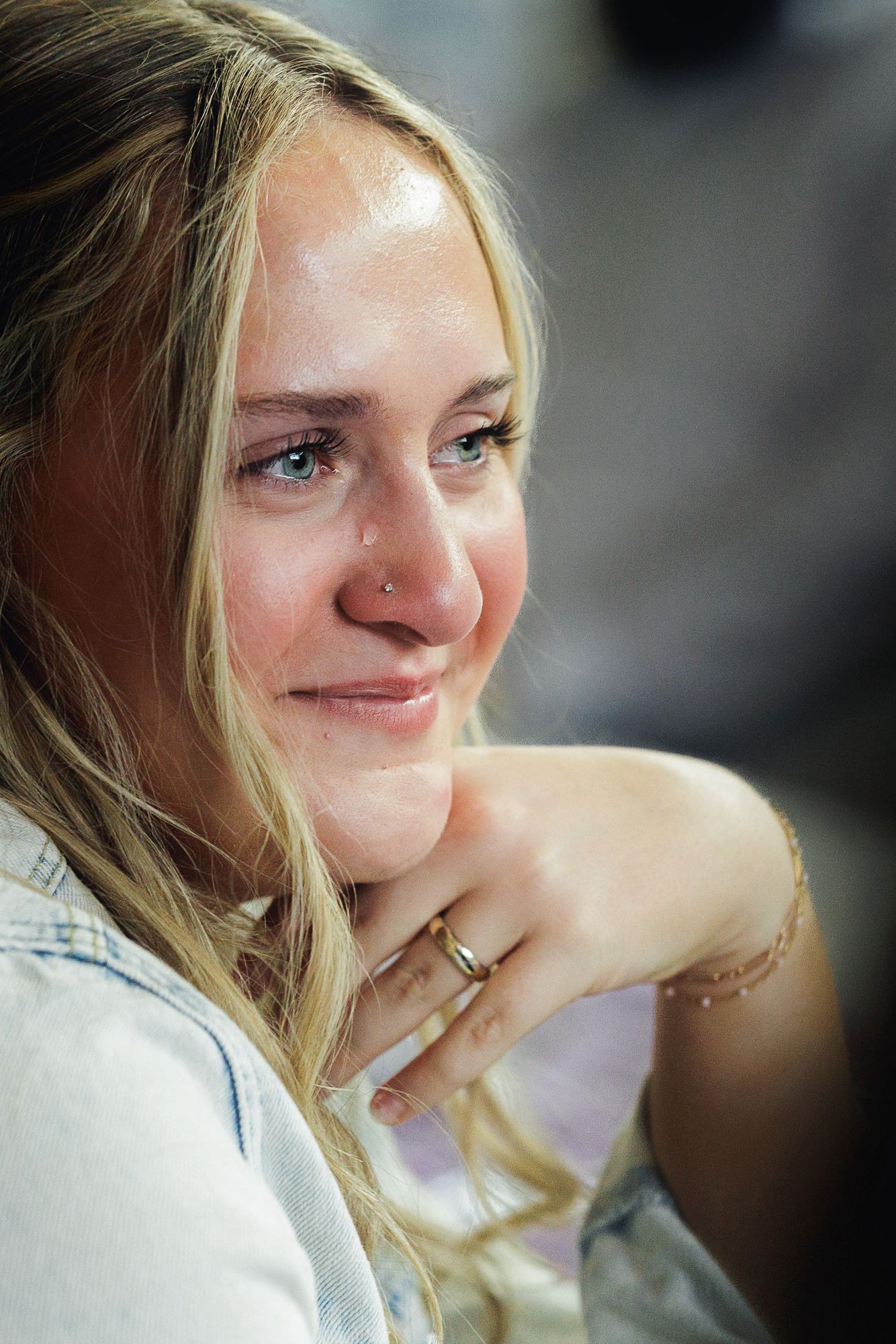 Close-up portrait of a smiling young blonde woman with blue eyes, wearing a ring and delicate necklace.