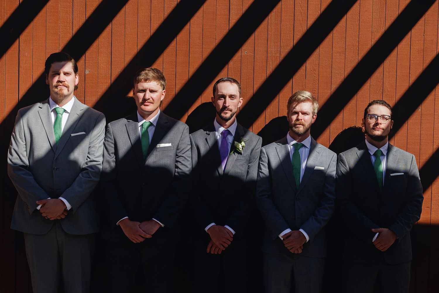 Five groomsmen in gray suits with green ties pose together outdoors against a striped wooden wall.