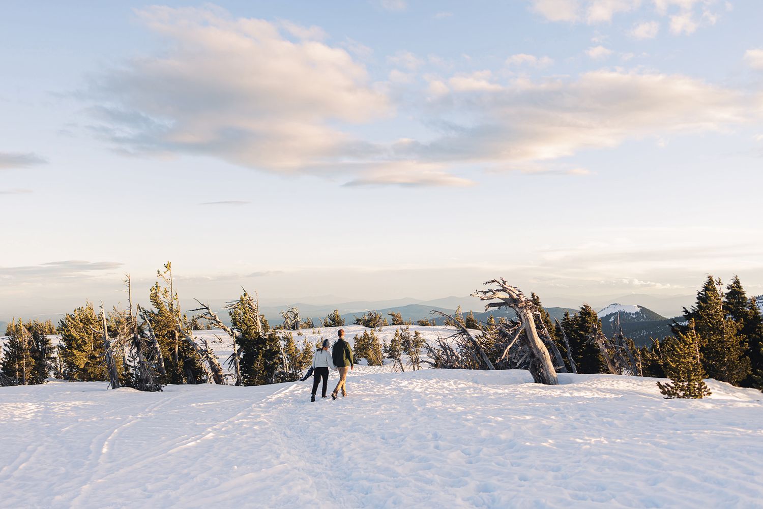 Two people walk through a snowy mountain landscape with evergreen trees and a ski lift under a pastel winter sky.