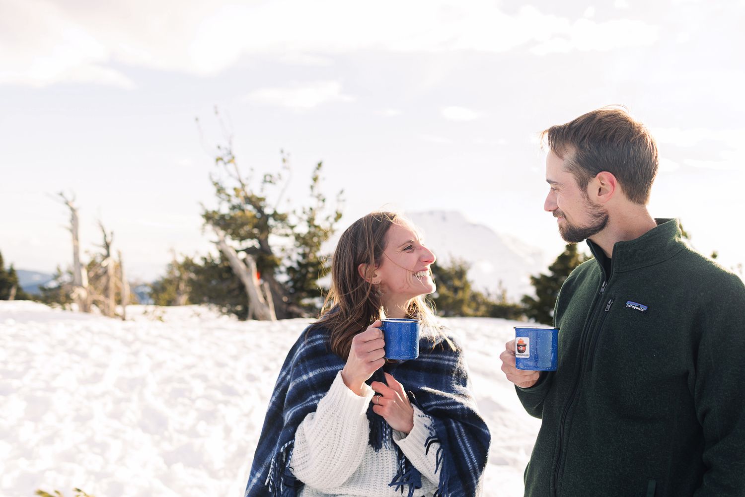 Couple smiling and holding blue mugs in a snowy winter landscape, dressed in cozy outdoor clothing.