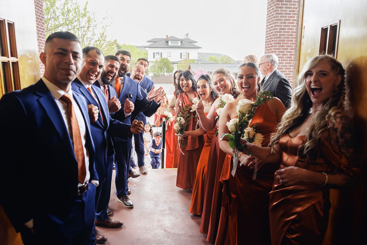 Wedding party in rust orange bridesmaid dresses and navy suits celebrating joyfully in a church doorway.