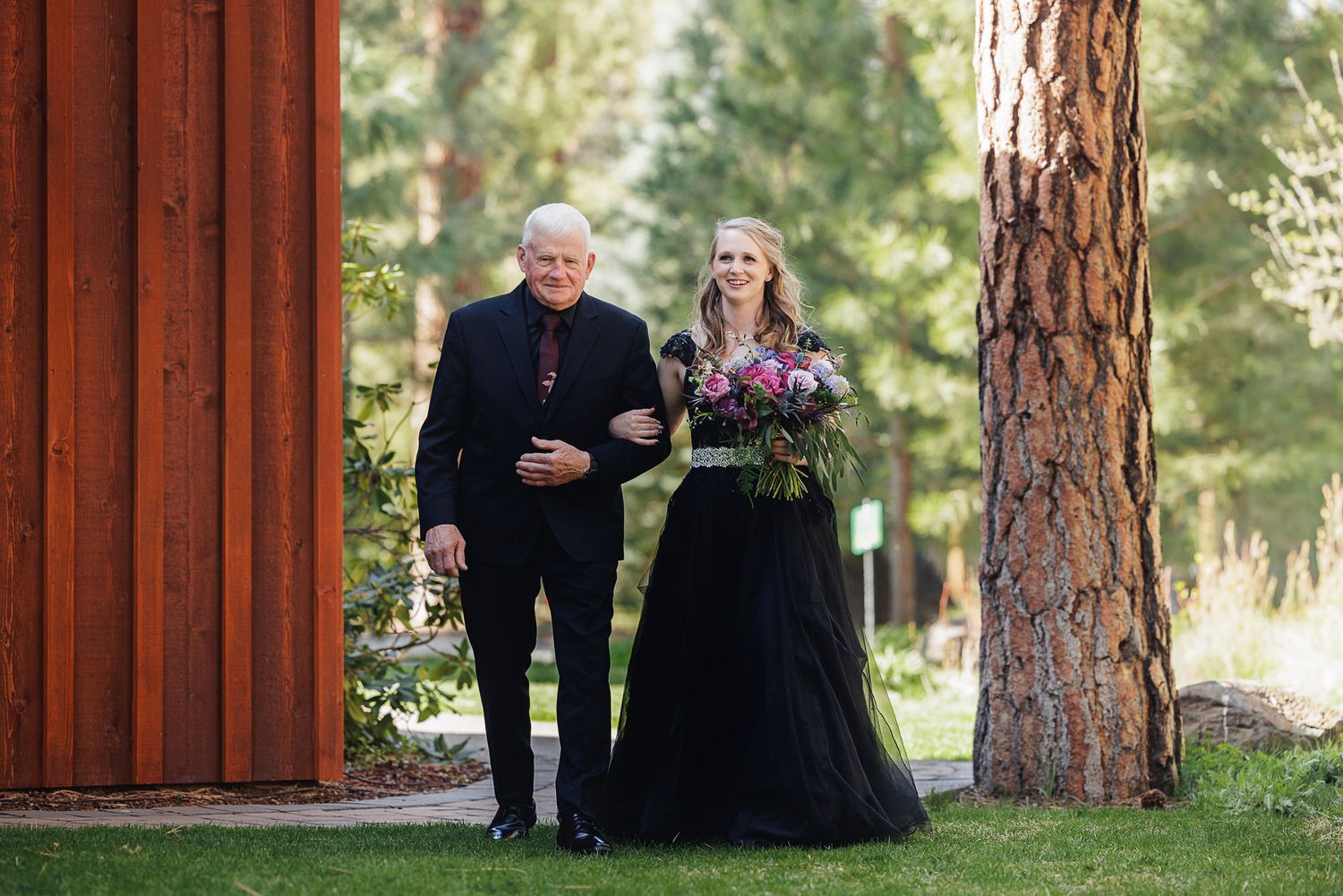 Bride in black wedding gown walks with older man through pine forest setting at outdoor wedding ceremony.