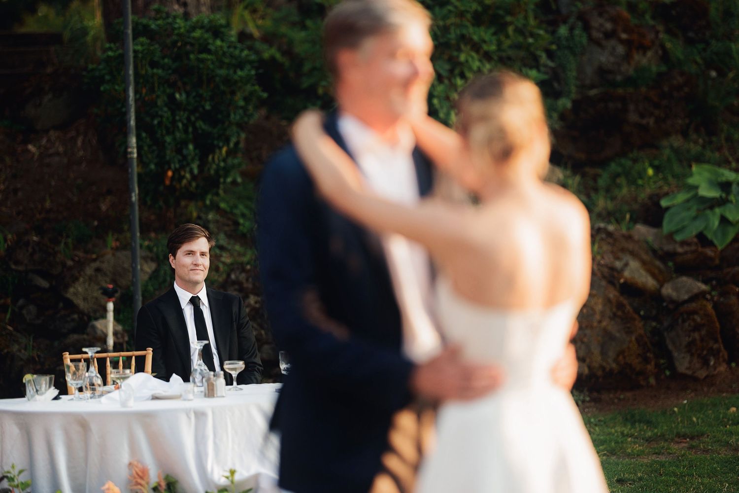 Blurred bride and groom share first dance while guest watches at outdoor wedding reception.