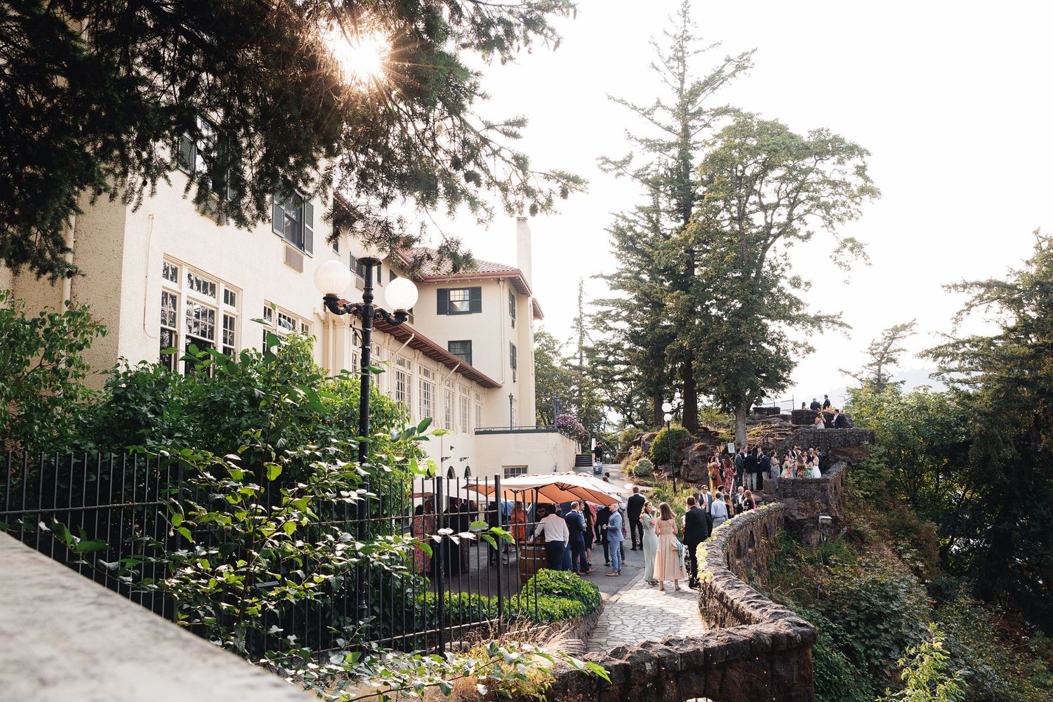 Guests gather outside an elegant villa surrounded by lush trees during a sunlit outdoor event.