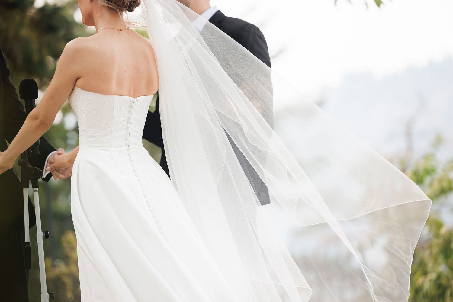 Bride in white strapless gown with flowing veil stands beside groom in black suit at outdoor wedding ceremony.
