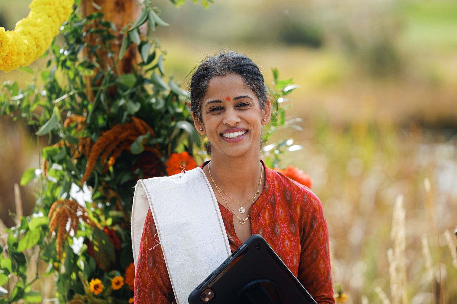 Smiling Indian woman in red saree holding clipboard outdoors among colorful flowers in a sunny field.