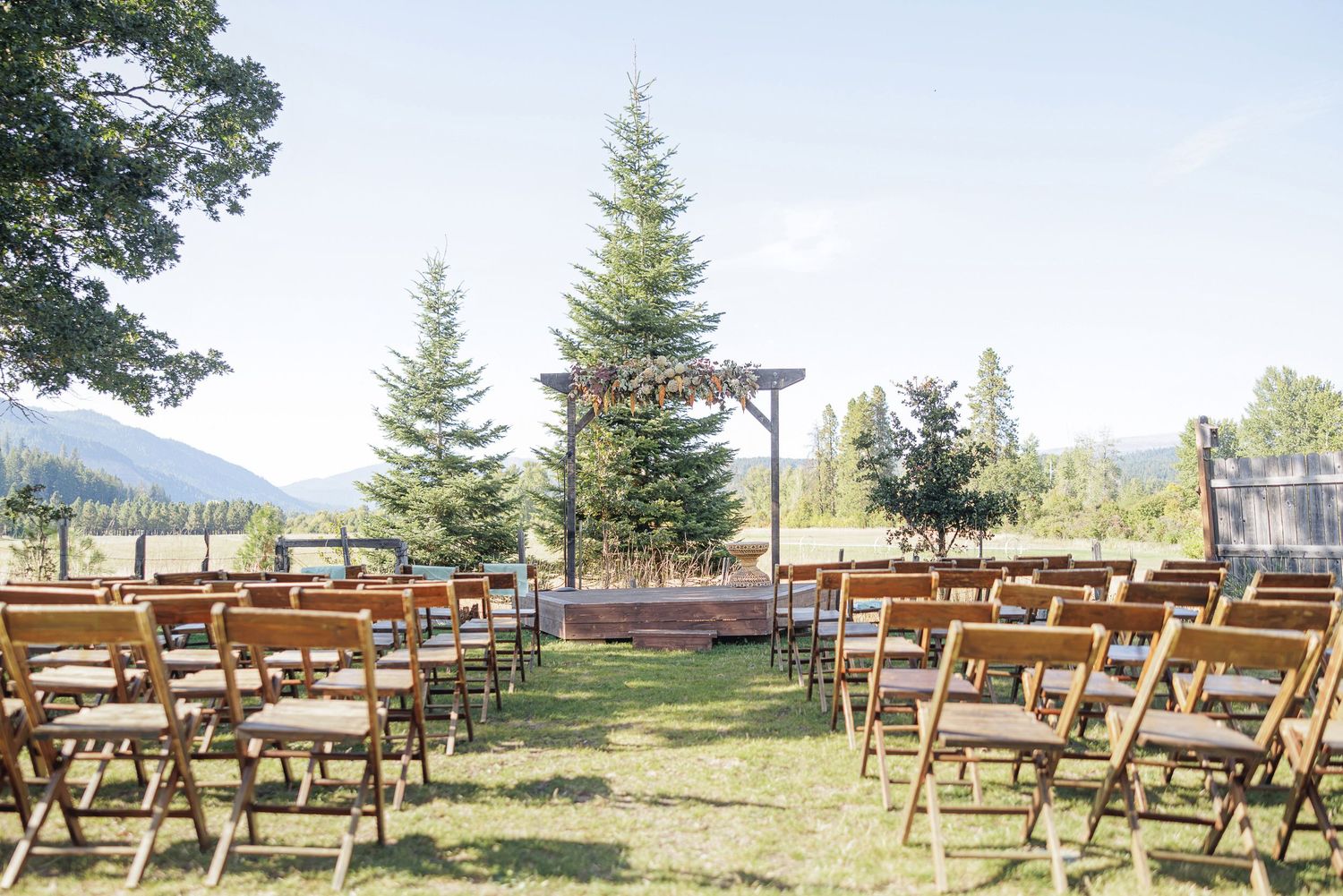 Outdoor wedding ceremony setup with wooden chairs, rustic arbor, tall pine trees, and mountain views on a sunny day.