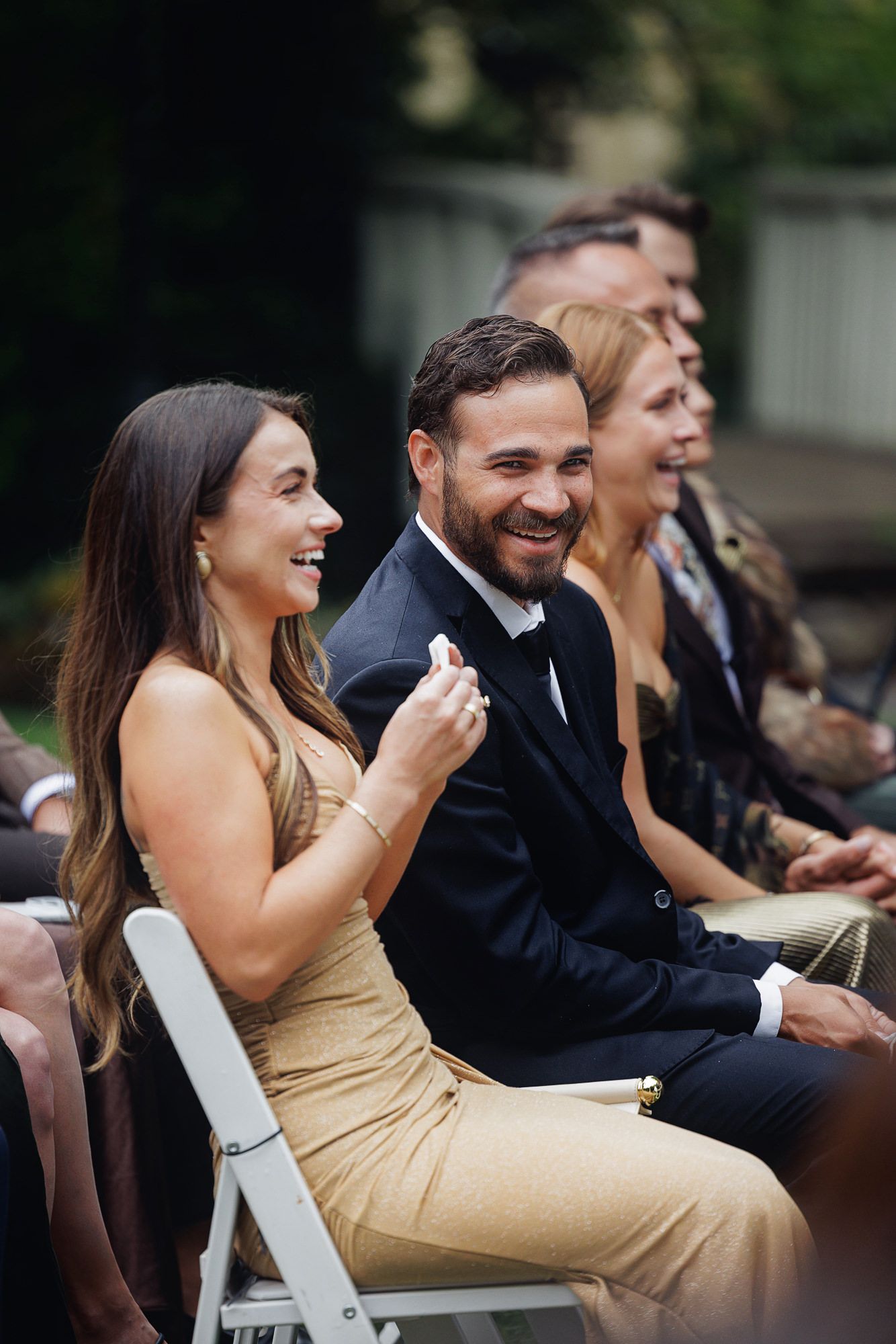 Smiling wedding guests seated outdoors, woman in gold dress and man in black suit enjoying the ceremony.