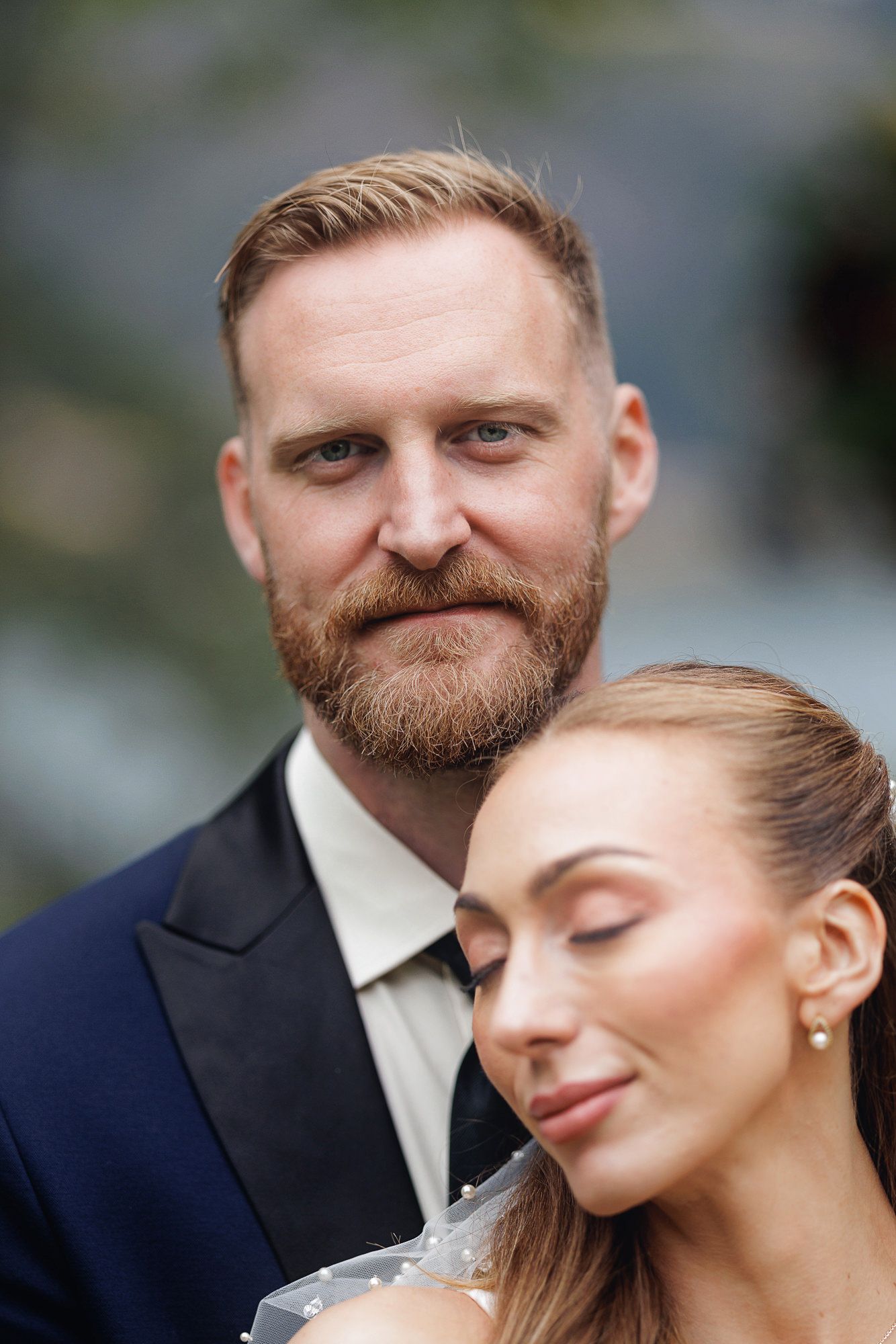 Bearded groom in navy suit gazes at camera while bride in white veil leans against him with eyes closed.