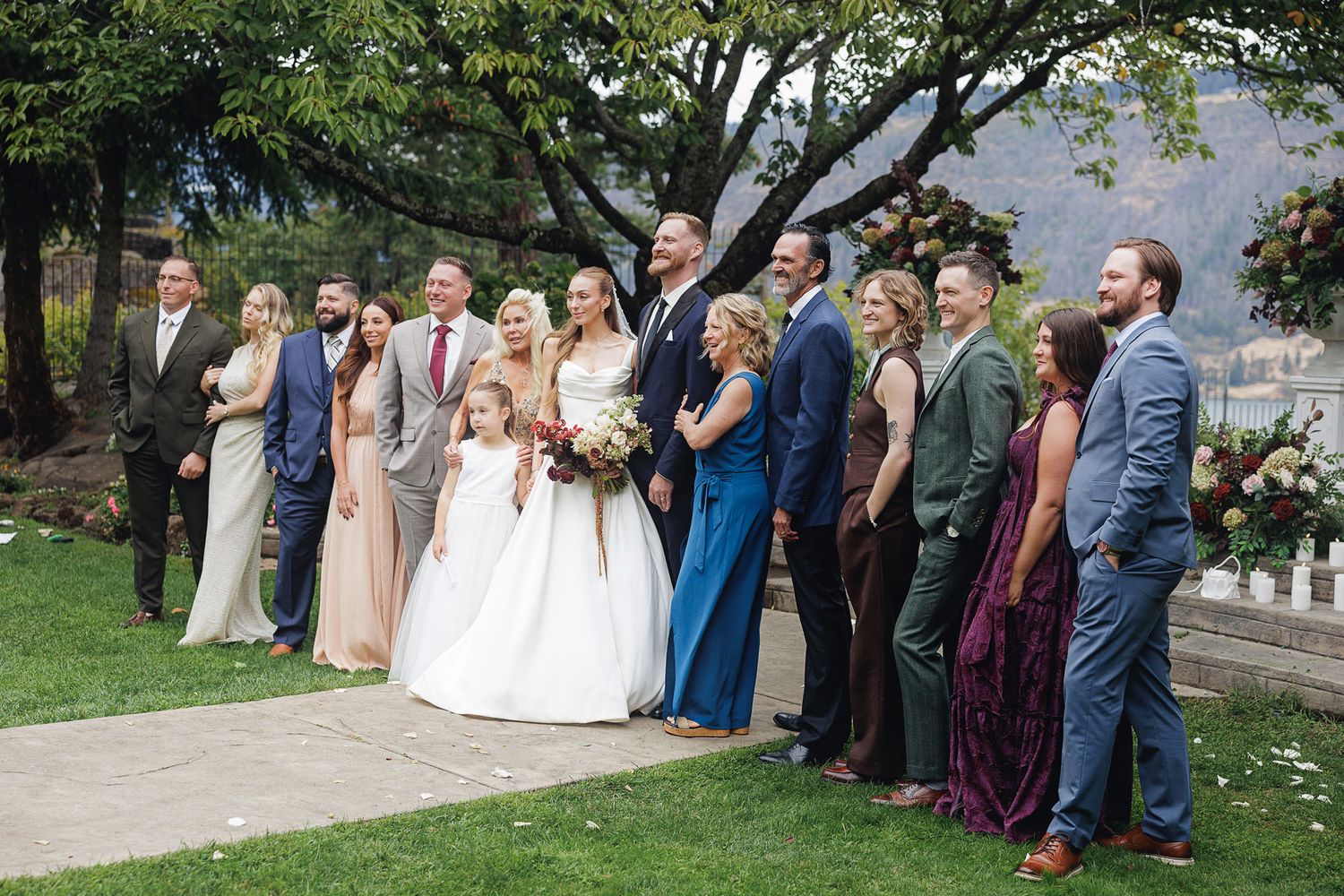 Wedding party poses outdoors on green lawn, bride in white gown surrounded by bridesmaids and groomsmen in colorful formal attire.